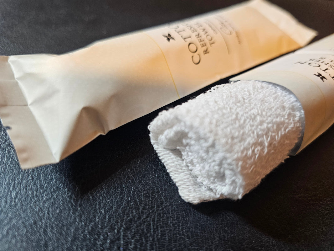 Two Cotton Refresher Towels in paper wrappers, one unrolled, on a dark counter